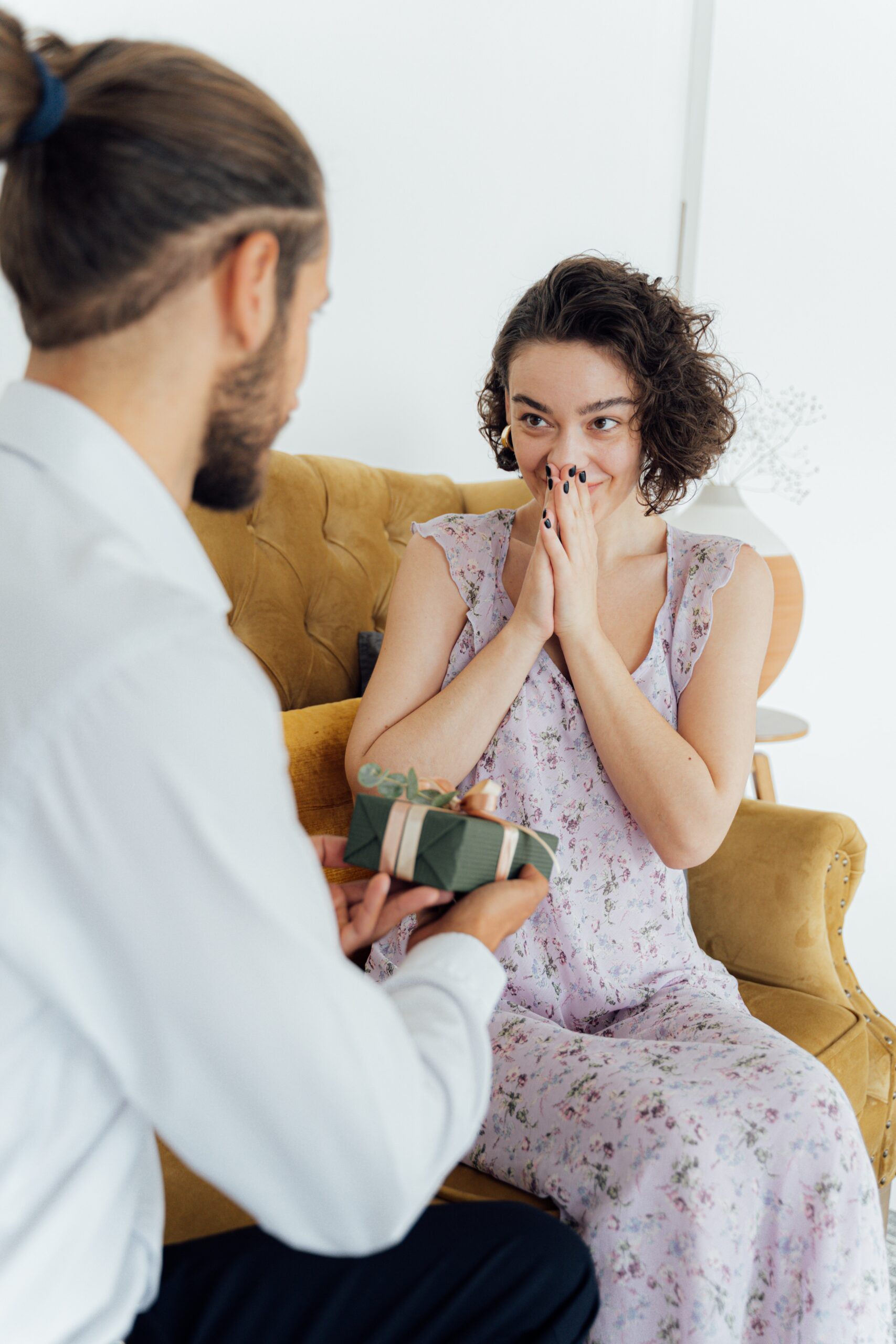 Image of a man presenting a woman with a gift box. The man's back is to the camera and the woman is facing the camera with her hands up to her face in surprise.