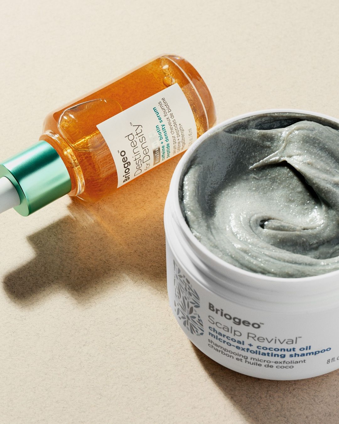 There are two hair products. The first, at the top, is in a clear dropper bottle. The contents of the products are golden. The other product is in a larger tub and is a grey-green in colour.