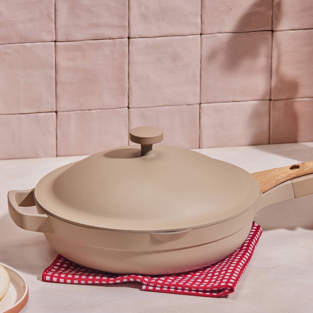 A neutral coloured pan with a matching lid and wooden handle is sitting on a red and white checkered cloth. The background is a pink textured subway tile.