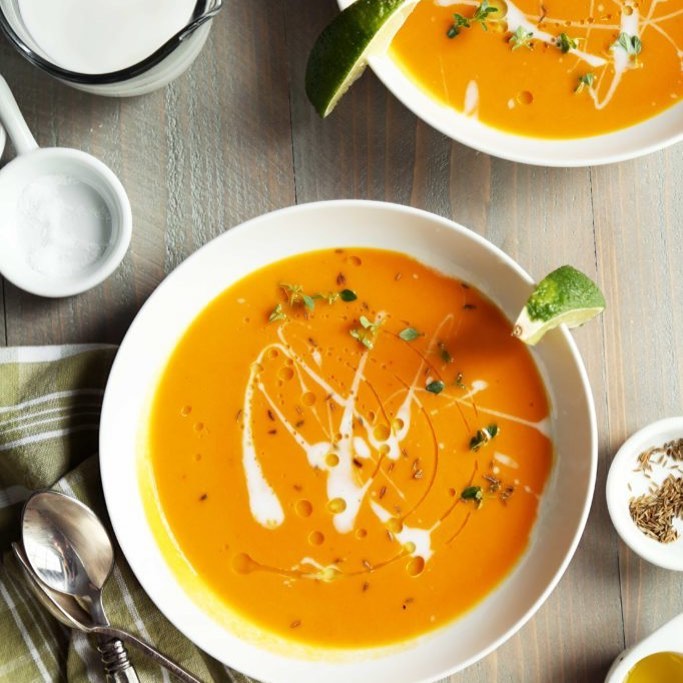 There are two bowls of an orange soup with white cream and a lime as a garnish. Beside the bowls is a green napkin with a spoon.