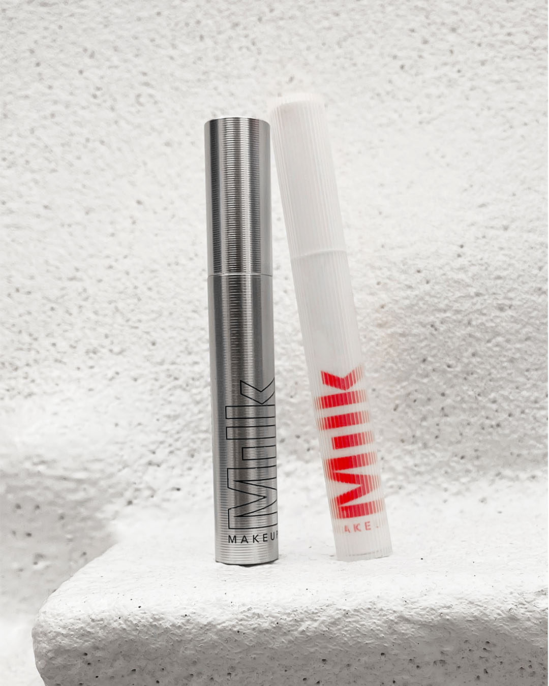 Two mascara products sitting upright against each other. One is silver and the other is white.