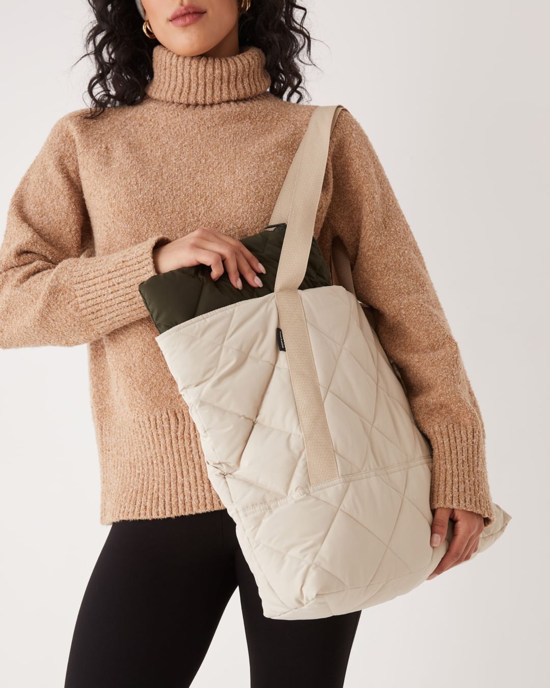 Model in a beige turtleneck and black pants carrying a large, puffy cream tote bag worn over her shoulder.